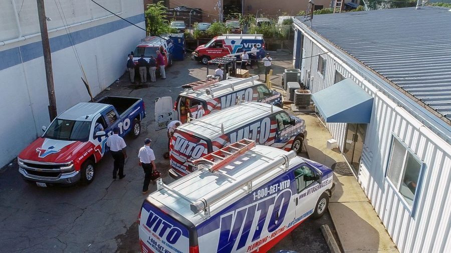 Aerial view of a service yard with multiple VITO-branded plumbing vans parked in a row beside a white building, workers standing nearby.