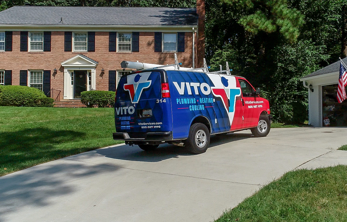 Colorful VITO plumbing van parked on a residential driveway in front of a brick house with green lawn and trees behind it.