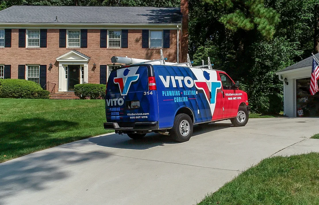 Colorful VITO plumbing van parked on a residential driveway in front of a brick house with green lawn and trees behind it.